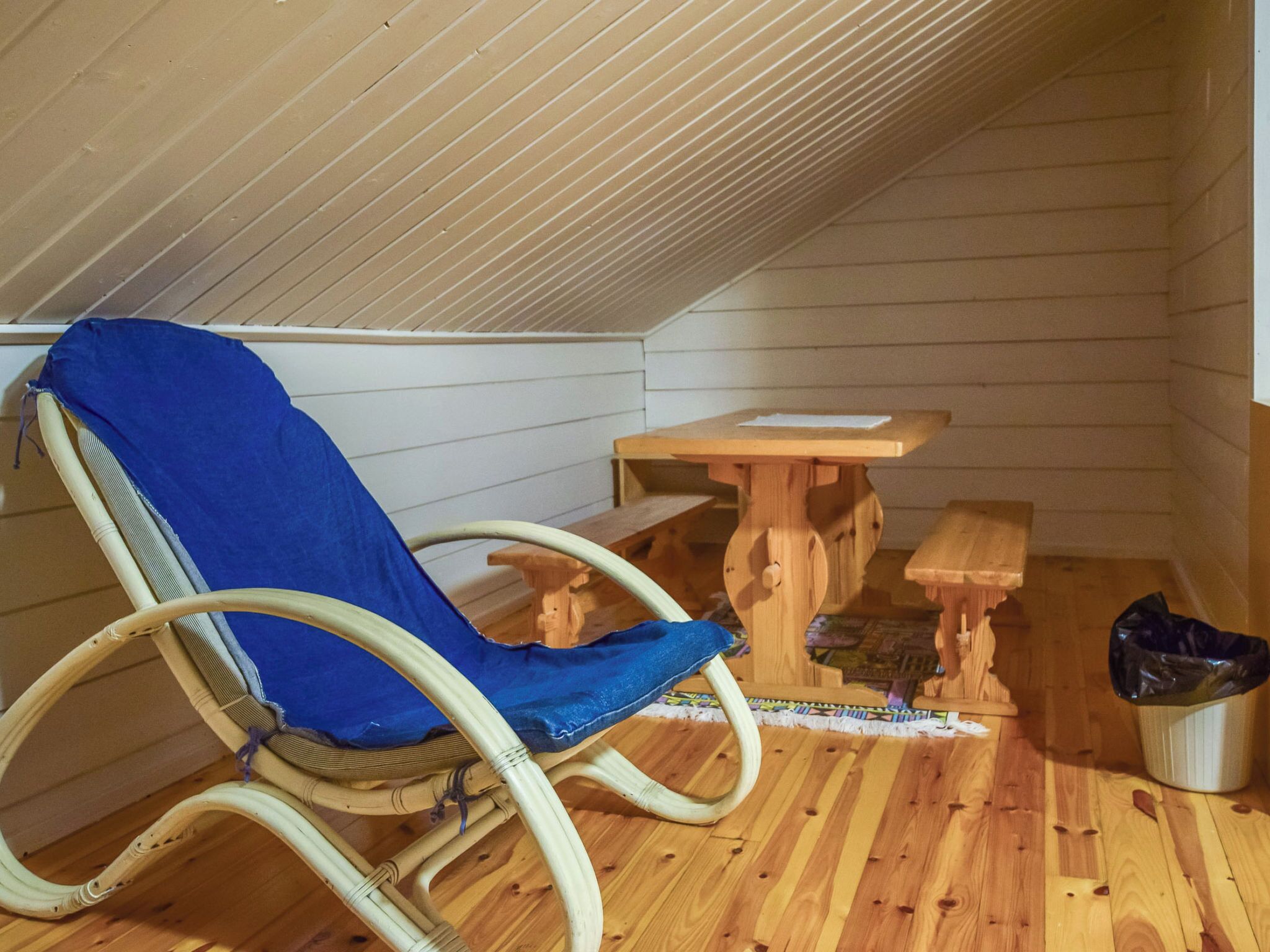 A small wooden room with a rocking chair, a wooden table, and a trash bin.