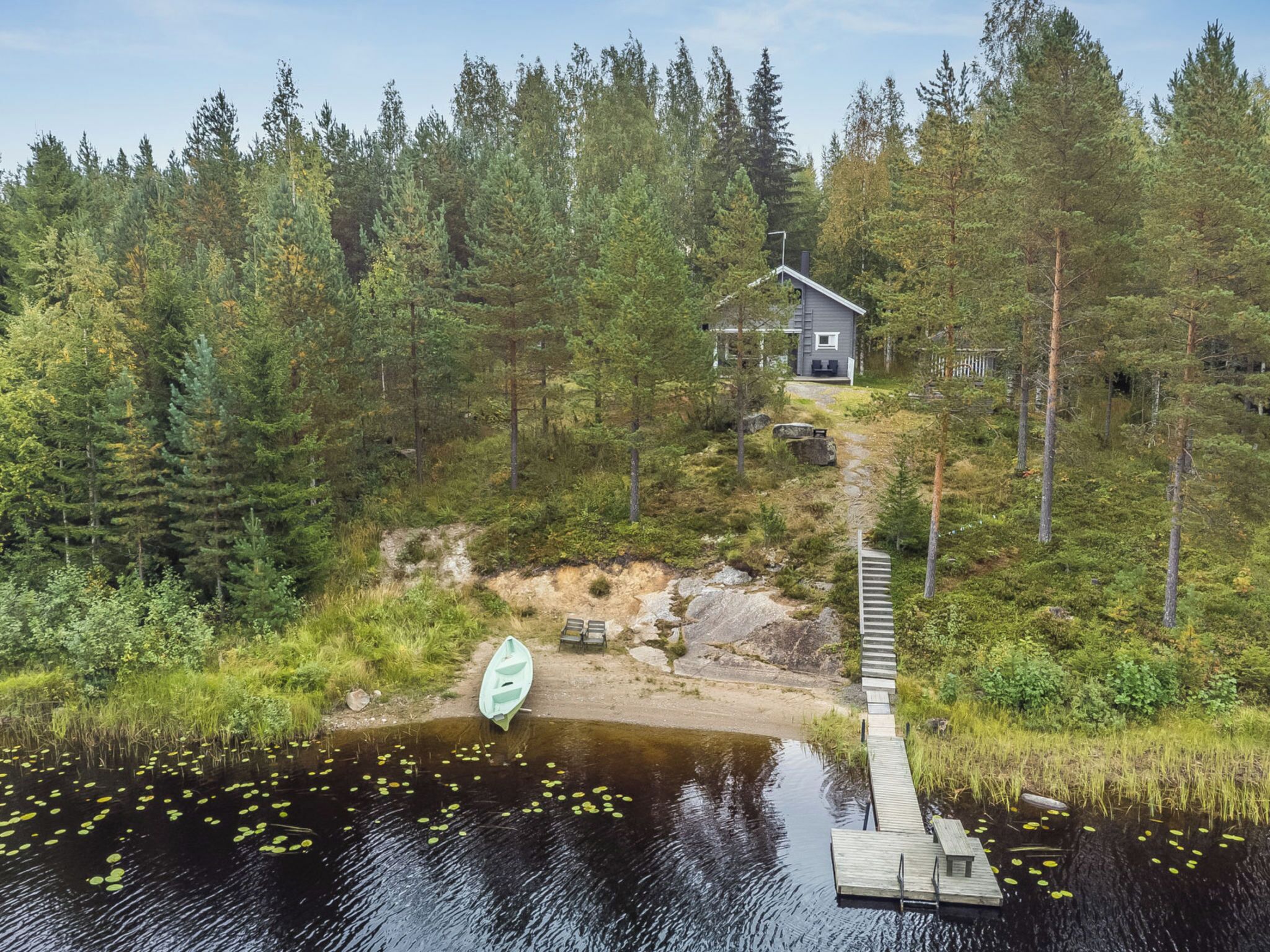 Une cabane entourée d’une forêt dense, une jetée s’avançant dans un lac parsemé de nénuphars, et un petit bateau.