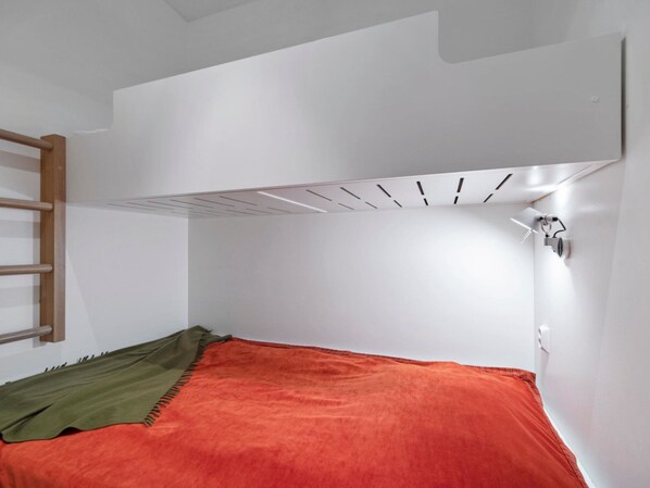 A small, compact room with a bunk bed, a wooden ladder, a white ceiling with a vent, and a wall-mounted light fixture.