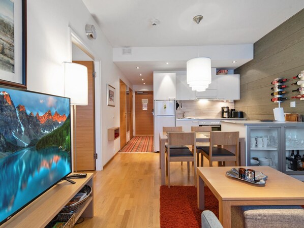 A modern hotel room with a wooden floor, a dining area with a table and chairs, a kitchen with white cabinets, and a mounted TV displaying a scenic mountain view.