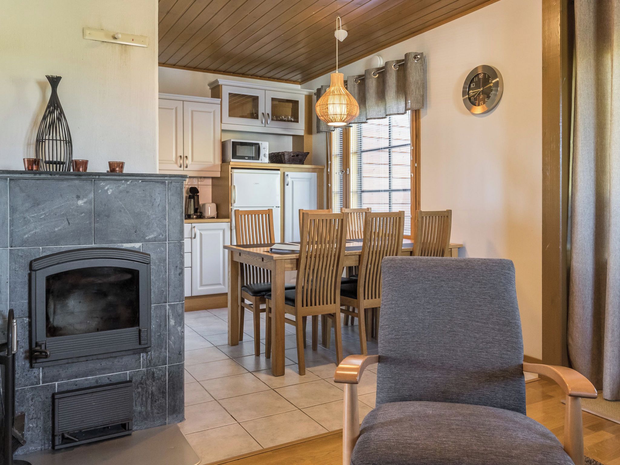 A cozy living room with a fireplace, wooden dining table, and chairs, a kitchen with cabinets and appliances, and a large window with curtains.