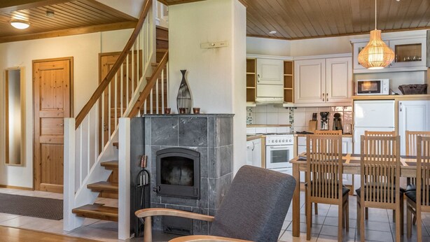 A cozy living room with a fireplace, wooden staircase, and a dining area with wooden chairs and a table. - Alppitähti by Interhome (Nilsiä)