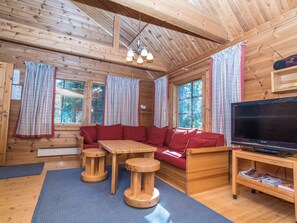 L’intérieur d’un chalet en bois, avec un canapé, deux tables en bois, un téléviseur et une fenêtre avec des rideaux.