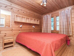 Habitación de cabaña de madera con una cama, una mesita de noche, un estante, una ventana con cortinas y un aplique de techo.