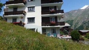 A multi-story hotel with balconies, a mountainous backdrop, and a garden area with outdoor seating. - Apartment Adler by Interhome (Saas-Fee)