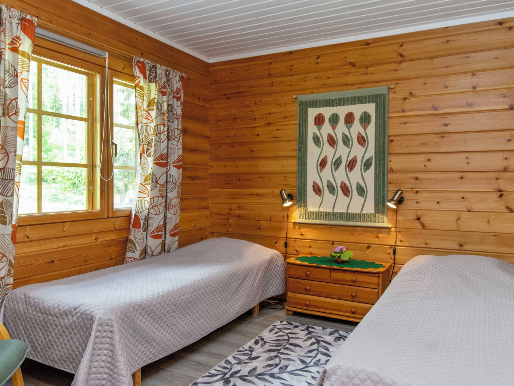 Une chambre de style cabane avec deux lits, des murs en bois, une fenêtre avec des rideaux, une lampe fixée au mur et une petite table avec une plante.