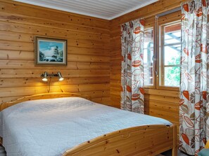 A wooden cabin bedroom with a bed, a painting on the wall, and curtains with a leaf pattern. - Vacation home 7152 by Interhome (Sulkava)