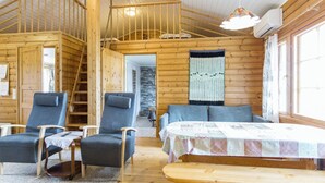 A cabin interior with a wooden staircase, two armchairs, a bed with a quilt, and a wooden table.