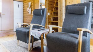 A cabin interior with two grey upholstered chairs, a wooden table, a white refrigerator, and a wooden ladder.