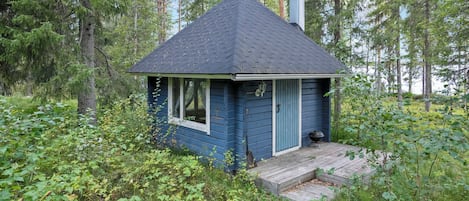 En liten stuga med ett mörkt tak, omgiven av tät grönska och träd.