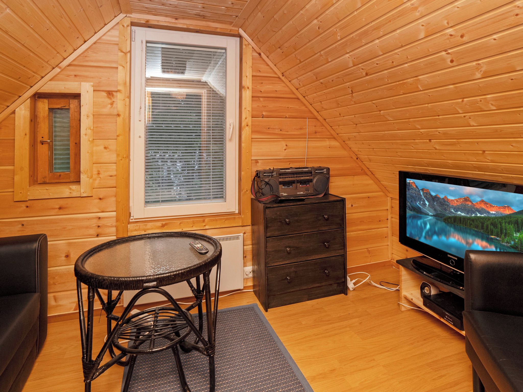 A cozy room with a wooden interior, a television, a small table with a chair, and a window with blinds.