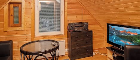 A cozy room with a wooden interior, a television, a small table with a chair, and a window with blinds.