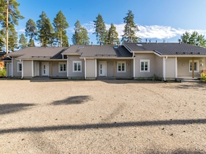A row of single-story buildings with a gravel driveway, surrounded by trees and a clear sky.