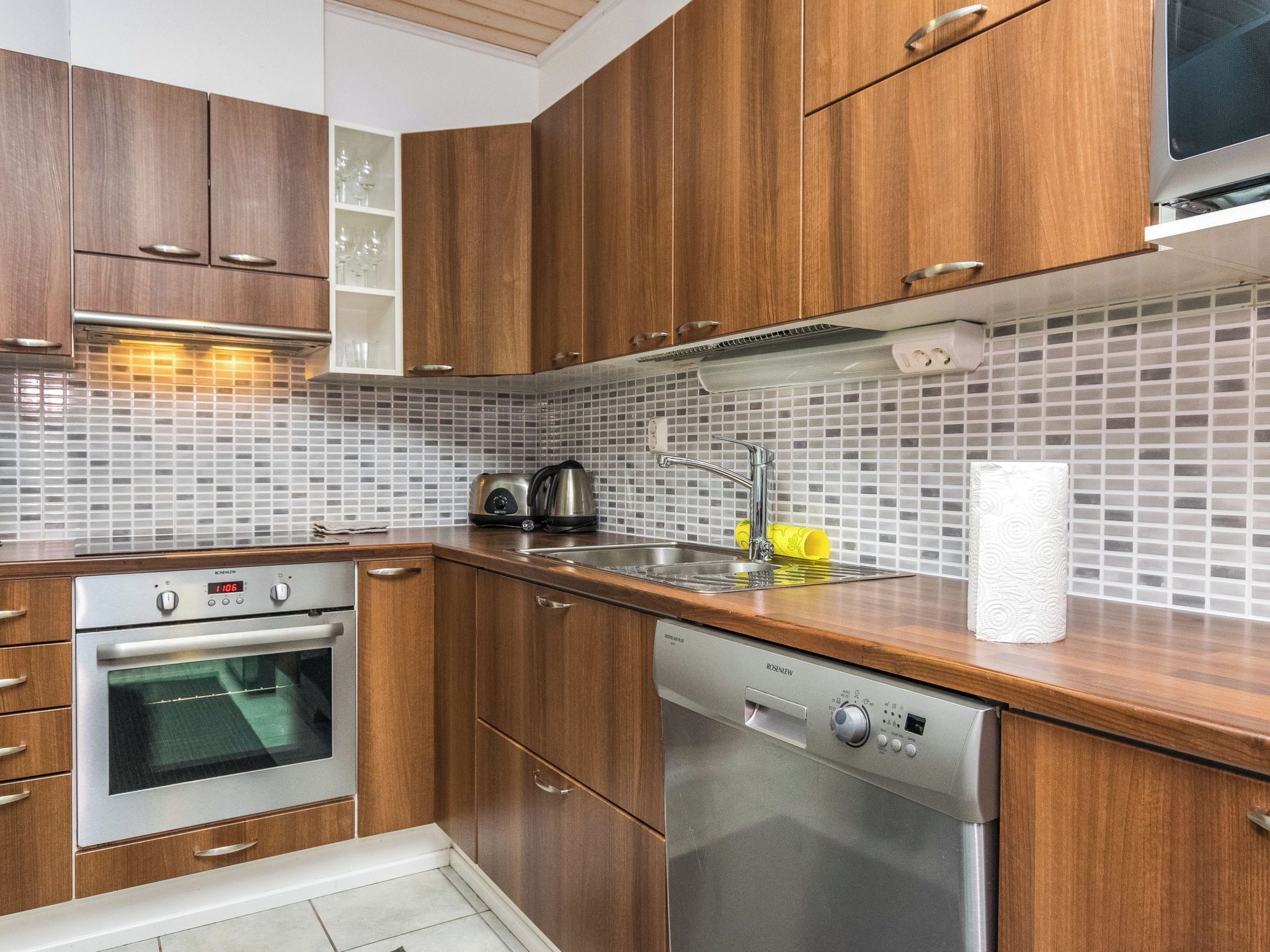 Eine moderne Küche mit Holzschränken, einem Edelstahlbackofen, einer Spülmaschine und einer gefliesten Spritzschutzwand.