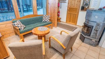 A cozy cabin interior with a sofa, chairs, a round wooden table, a fireplace, and a window overlooking a snowy landscape.
