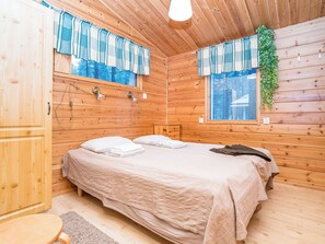 A wooden cabin room with a bed, two windows with curtains, a small table, and a wall-mounted light fixture. - Villa kuukkeli by Interhome (Kuusamo)