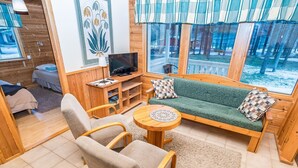 Ein gemütliches Wohnzimmer mit einer Couch, Sesseln, einem Fernseher und einem Fenster mit Vorhängen.
