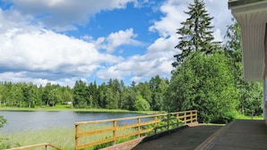 Une terrasse en bois mène à un lac, d’où l’on peut admirer une forêt dense et un bâtiment à l’extérieur blanc.