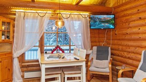 Ein gemütliches Blockhaushaus-Interieur mit Holzausstattung, einem Esstisch mit Stühlen, einem an der Wand montierten Fernseher und einem Fenster mit Vorhängen.