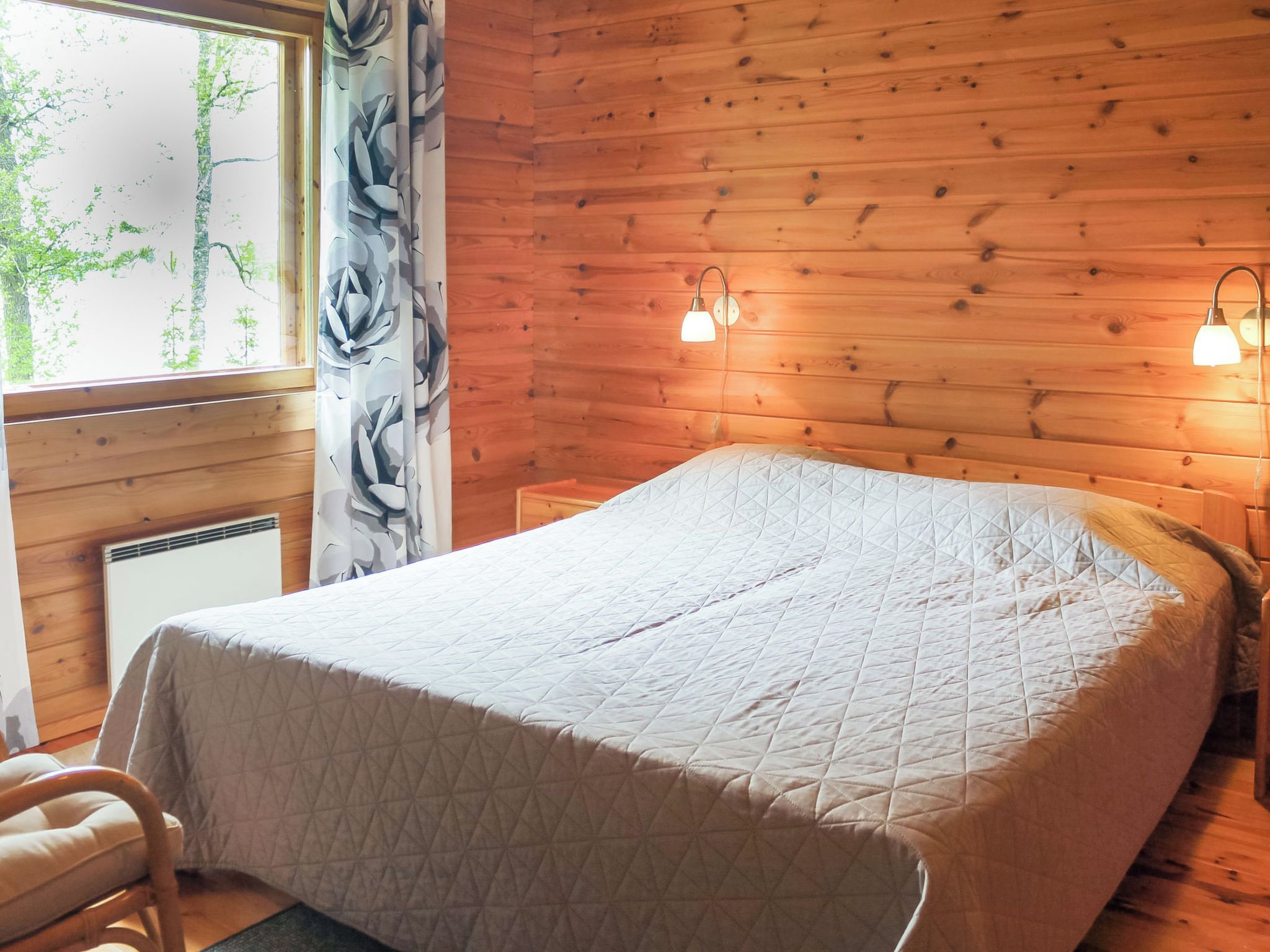 Quarto com parede de madeira, cama com colcha acolchoada, uma cadeira, janela com vista para árvores e duas luminárias fixadas na parede.