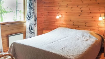 Quarto com parede de madeira, cama com colcha acolchoada, uma cadeira, janela com vista para árvores e duas luminárias fixadas na parede.