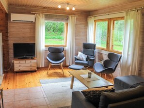 A cozy room with a TV, wooden furniture, and large windows offering a view of greenery.