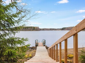 Una terraza de madera que conduce a un muelle sobre un lago tranquilo, rodeado de árboles y un cielo despejado.