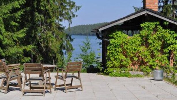 A wooden deck with chairs, a small building with a chimney, and a view of a lake.