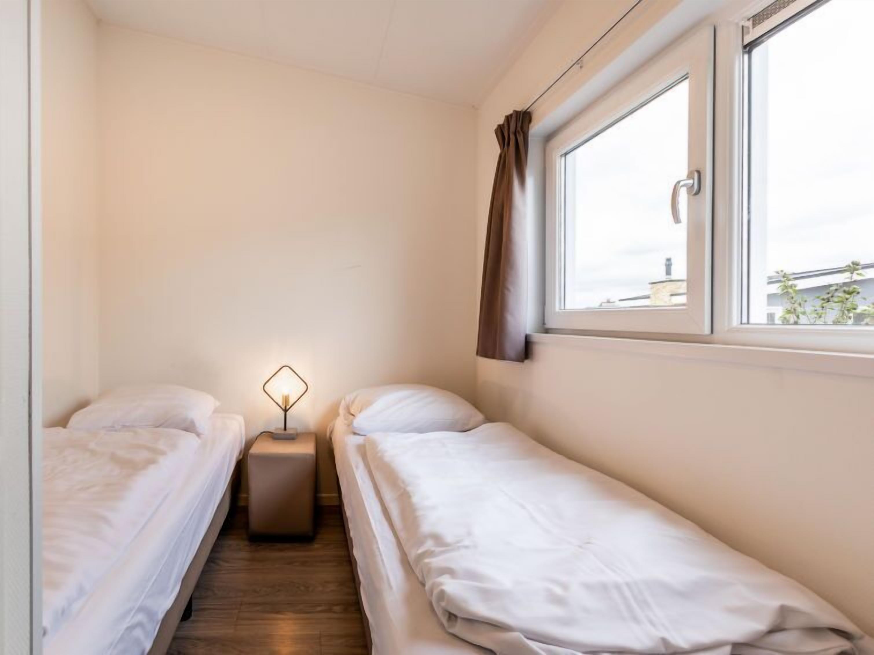 Una habitación pequeña y limpia con dos camas individuales, una mesita de noche, una lámpara y una ventana con cortinas.