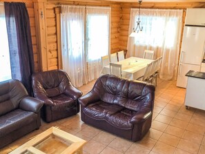 A log cabin interior with two leather sofas, a dining table with chairs, a chandelier, and a refrigerator.