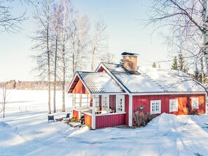 Una casa de madera roja con techo inclinado y chimenea, rodeada de nieve y árboles.