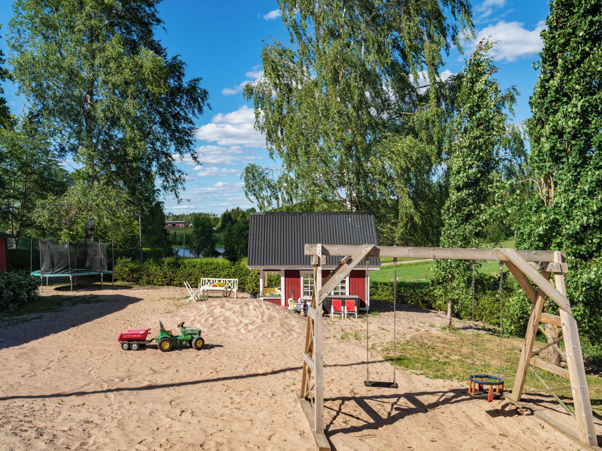 A sandy outdoor area with a swing set, a small red building, and a green tractor.