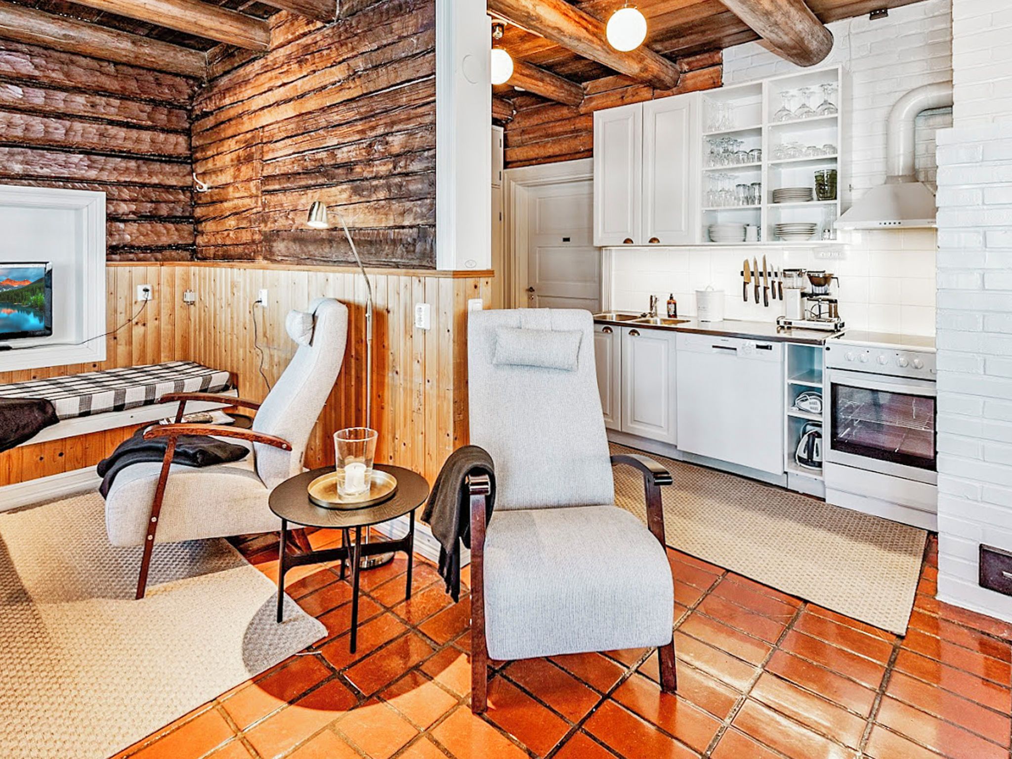 Een gezellig chaletinterieur met een houten wand, een eethoek met tafel en stoelen, en een keuken met witte kasten en een fornuis.