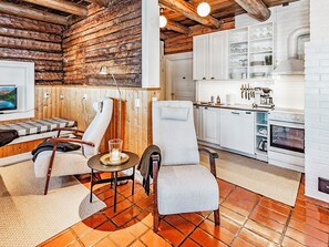 Interior acogedor de una cabaña con pared de troncos de madera, una zona de comedor con mesa y sillas, y una cocina con armarios blancos y una estufa.