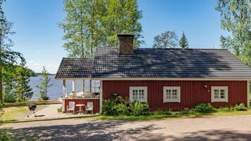A red wooden cabin with a tiled roof, a small porch with chairs, and a grill outside.