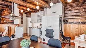 Un interior rústico con una mesa de comedor de madera, sillas, una planta en maceta y una cocina con armarios blancos y una chimenea de piedra.