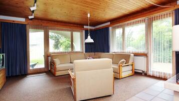 Ein geräumiges Wohnzimmer mit Holzbalkendecke, großen Fenstern, einem beigefarbenen Sofa und einem Fernseher.