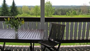 Um deck de madeira com uma mesa e uma cadeira, com vista para uma paisagem verde com árvores e céu claro.