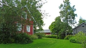 Uma casa de tijolos vermelhos com janela branca, cercada por vegetação exuberante e um gramado bem cuidado.