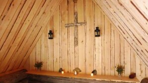Interior em madeira com uma cruz na parede, duas lanternas e um banco com plantas pequenas.
