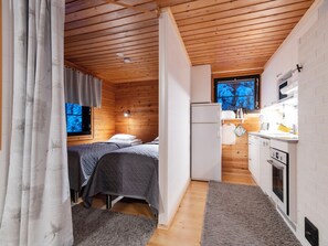 Ein gemütliches Blockhaus mit Bett, Kühlschrank, einer kleinen Küche und einem Fenster mit Blick auf Bäume.