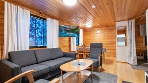 Interior acogedor de una cabaña con sofá, sillas, una mesa redonda y un amplio ventanal con vistas a árboles.