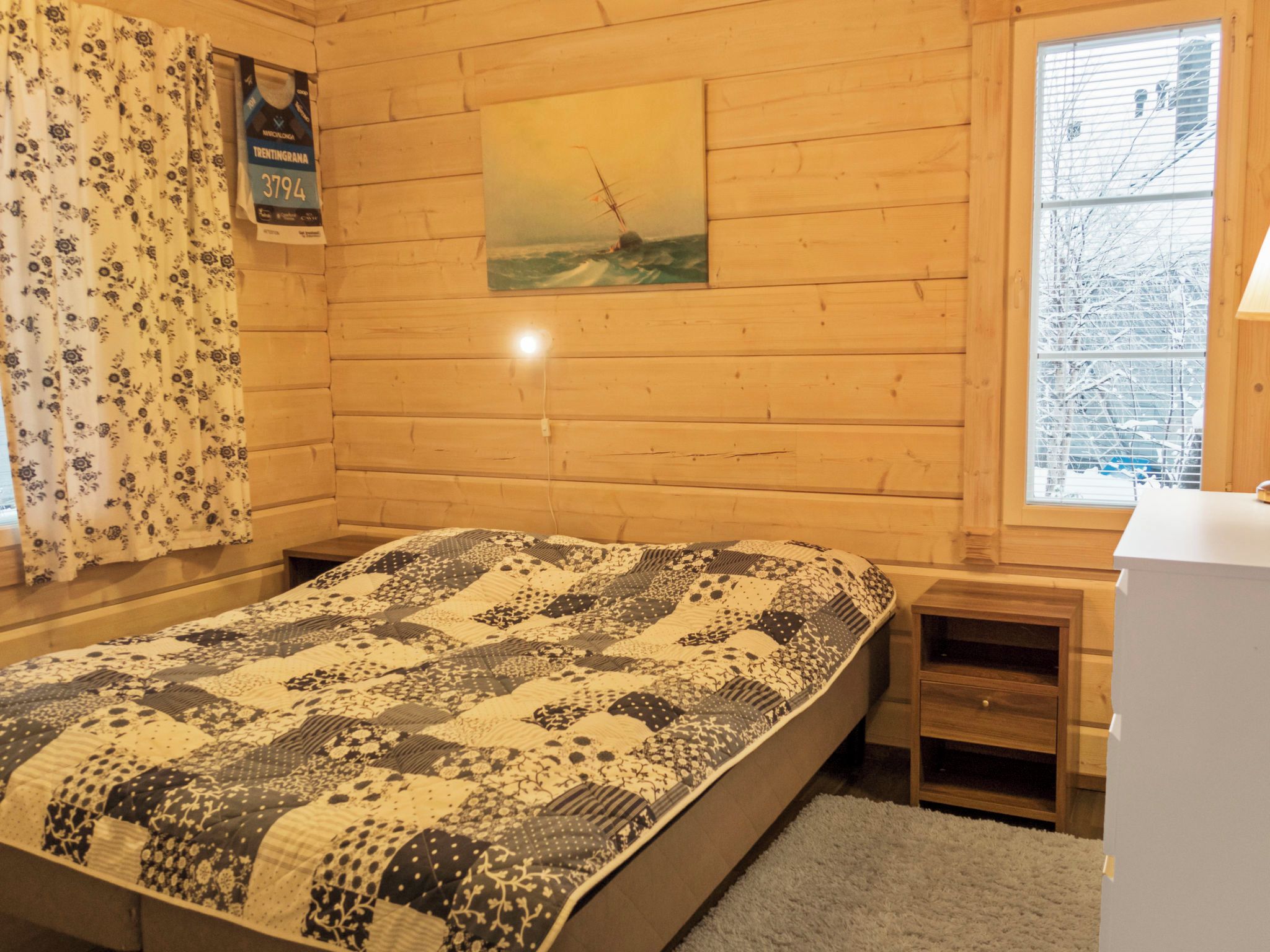 Een houten kamer met een bed, een klein nachtkastje, een raam met uitzicht op sneeuwbedekte bomen en een schilderij aan de muur.