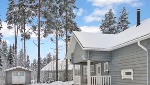 Une cabane à deux étages, dotée d’un porche et d’un petit abri, entourée d’arbres couverts de neige.