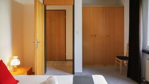 A hotel room with a bed, a red pillow, a white towel, a nightstand with a lamp, a wooden wardrobe, and a chair.