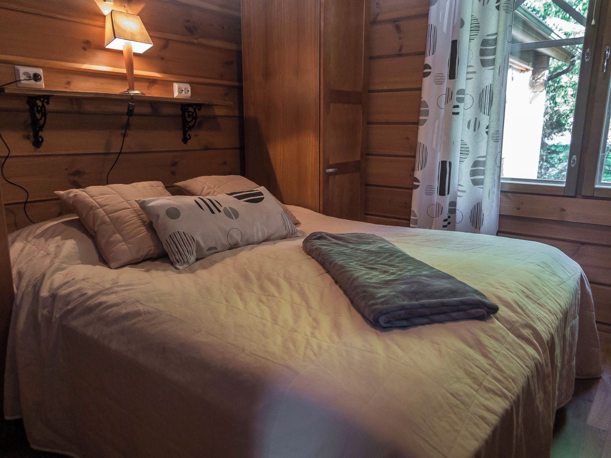 Ein Zimmer mit Holzboden, einem Bett, einer Wandleuchte und einem Fenster mit Vorhängen.