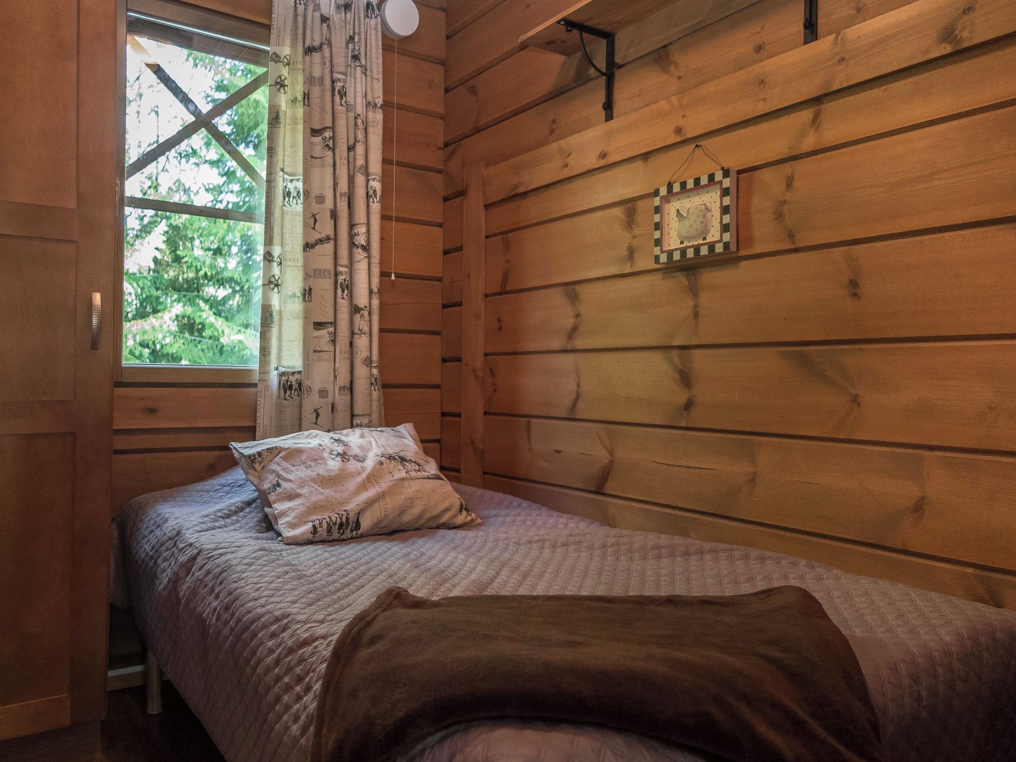 Ein Zimmer in einer Holzhütte mit einem Bett, einem Vorhangfenster und einer an der Wand befestigten Uhr.