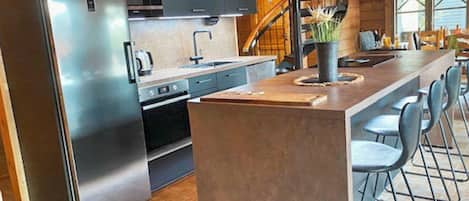 Cozinha moderna com geladeira de aço inoxidável, micro-ondas e forno embutido. Há uma ilha grande com banquetas e uma planta em vaso.