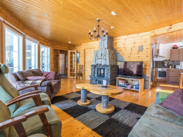 A cozy log cabin interior with a stone fireplace, wooden furniture, and a kitchen area.