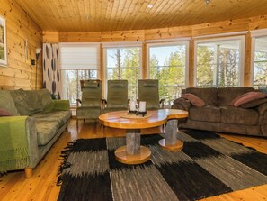 L’intérieur chaleureux d’un chalet avec un plafond en bois, deux canapés, une table ronde en bois et de grandes fenêtres offrant une vue sur les arbres.